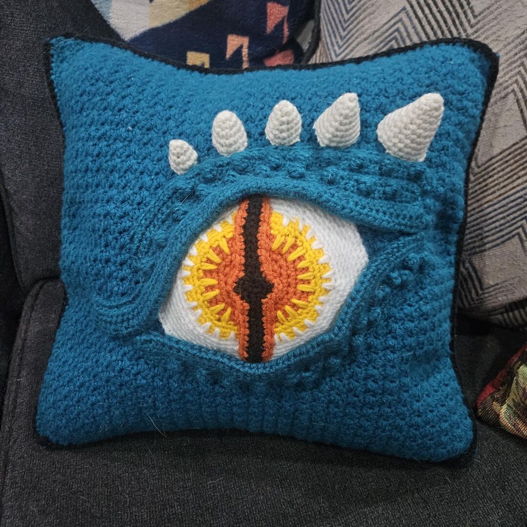 Blue crochet pillow with a dragon eye design on a dark surface